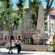 People sit by a fountain near trees, buildings and storefronts
